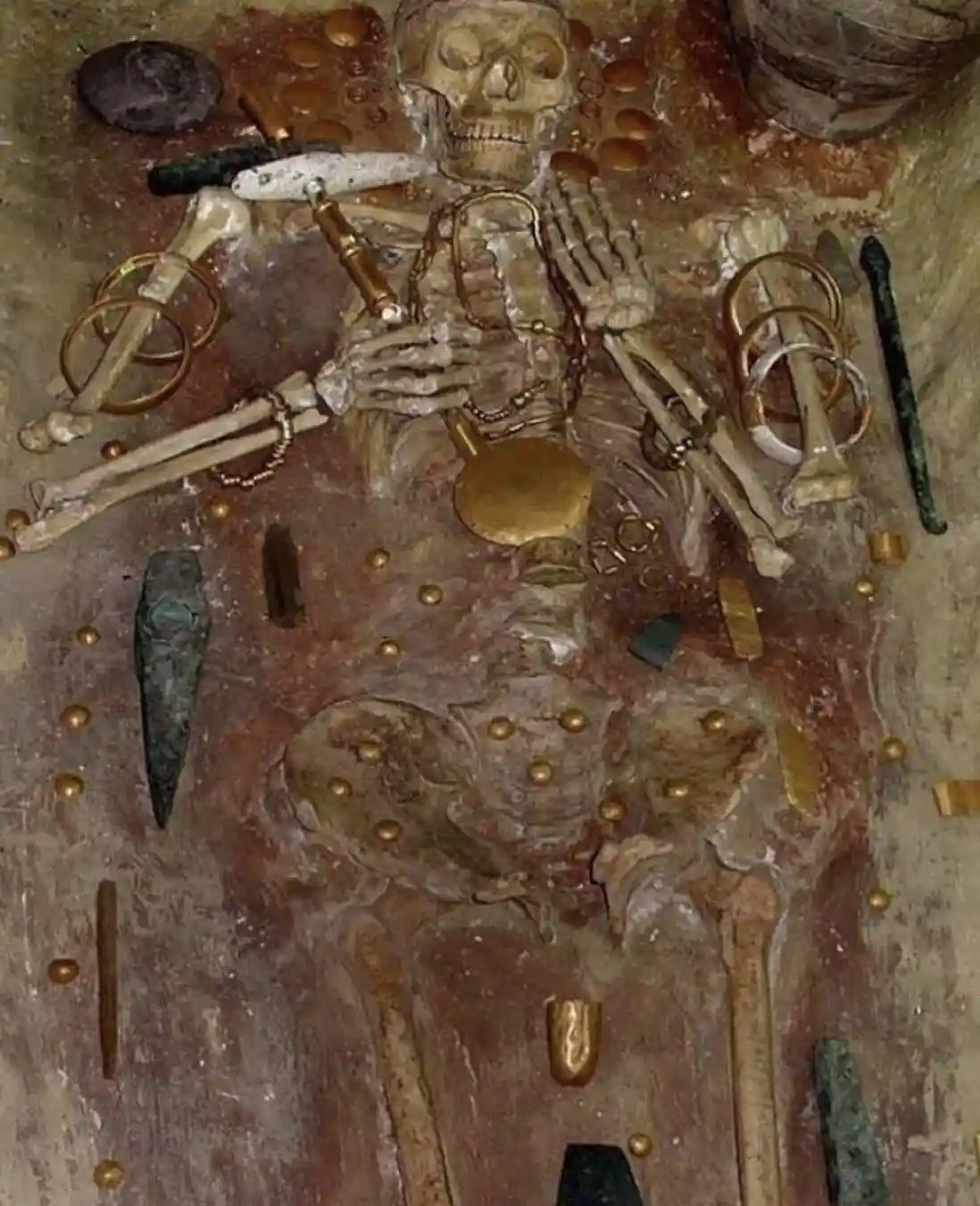 An ancient human skeleton lies embedded in earth, adorned with numerous golden coins, rings, and other metallic artifacts…