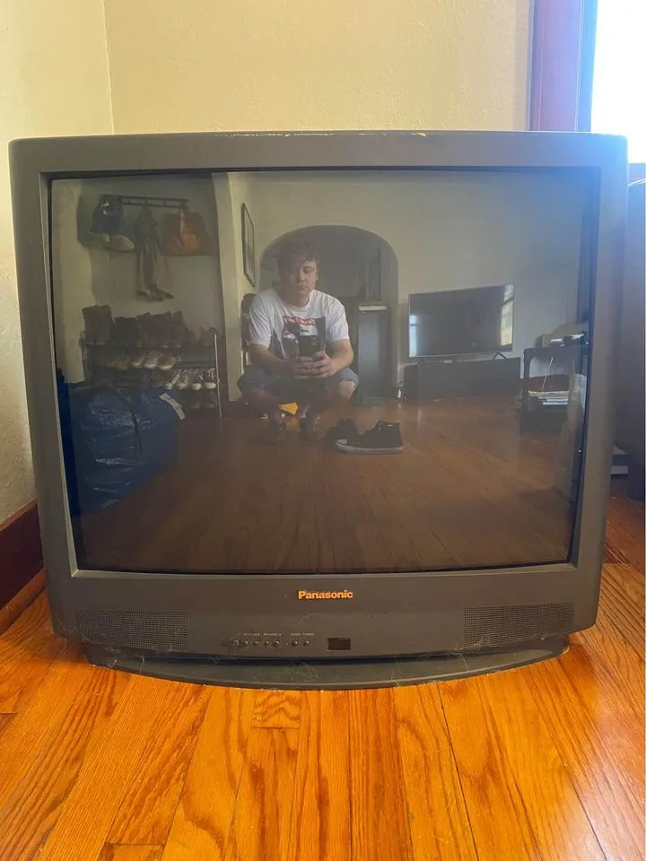 A person is crouching on a wooden floor, holding a phone, reflected in the screen of an old Panasonic CRT television. The…