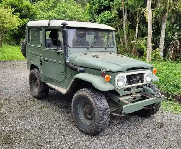 A green, rugged, vintage Toyota Land Cruiser Bandeirante is parked on a gravel path surrounded by lush green tropical…