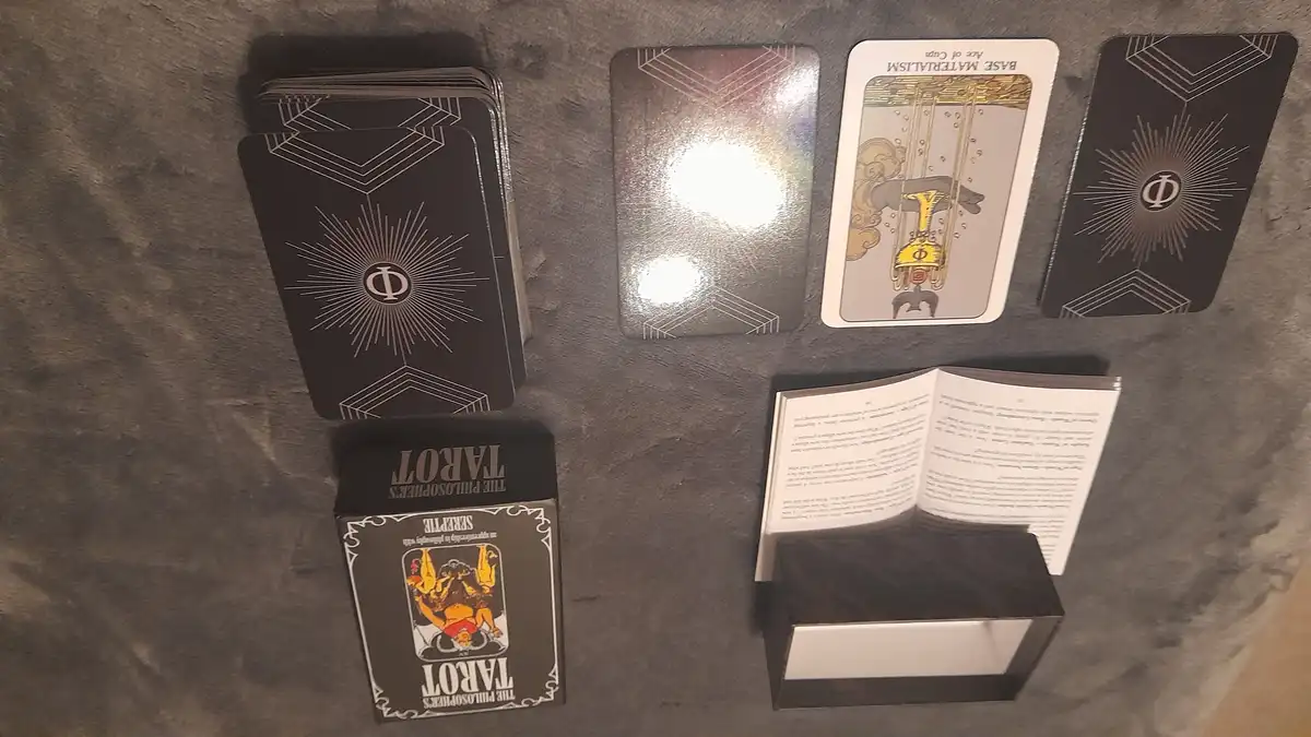 A spread of tarot cards, including a central card labeled 'BASE MATERIALISM' with an illustration of a hand holding a golden…