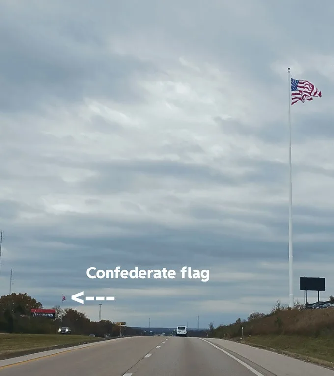 A highway scene under an overcast sky, featuring a tall flagpole with an American flag waving on the right. A white arrow…