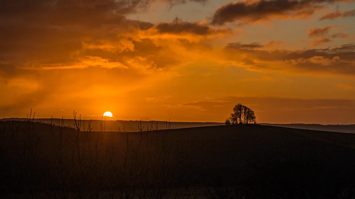 A serene landscape photo of a sunset with the sun low on the horizon, casting orange and yellow light across clouds…