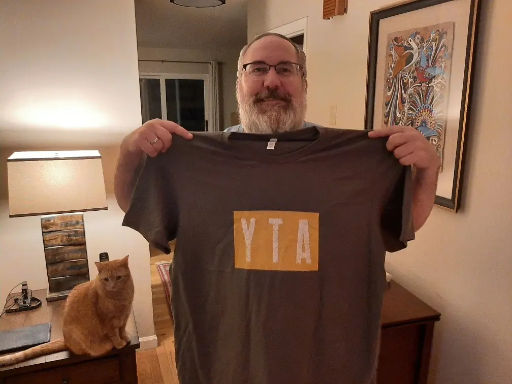 A man with glasses and a beard holds up a dark gray t-shirt with a yellow rectangle containing the letters 'YTA' in white…