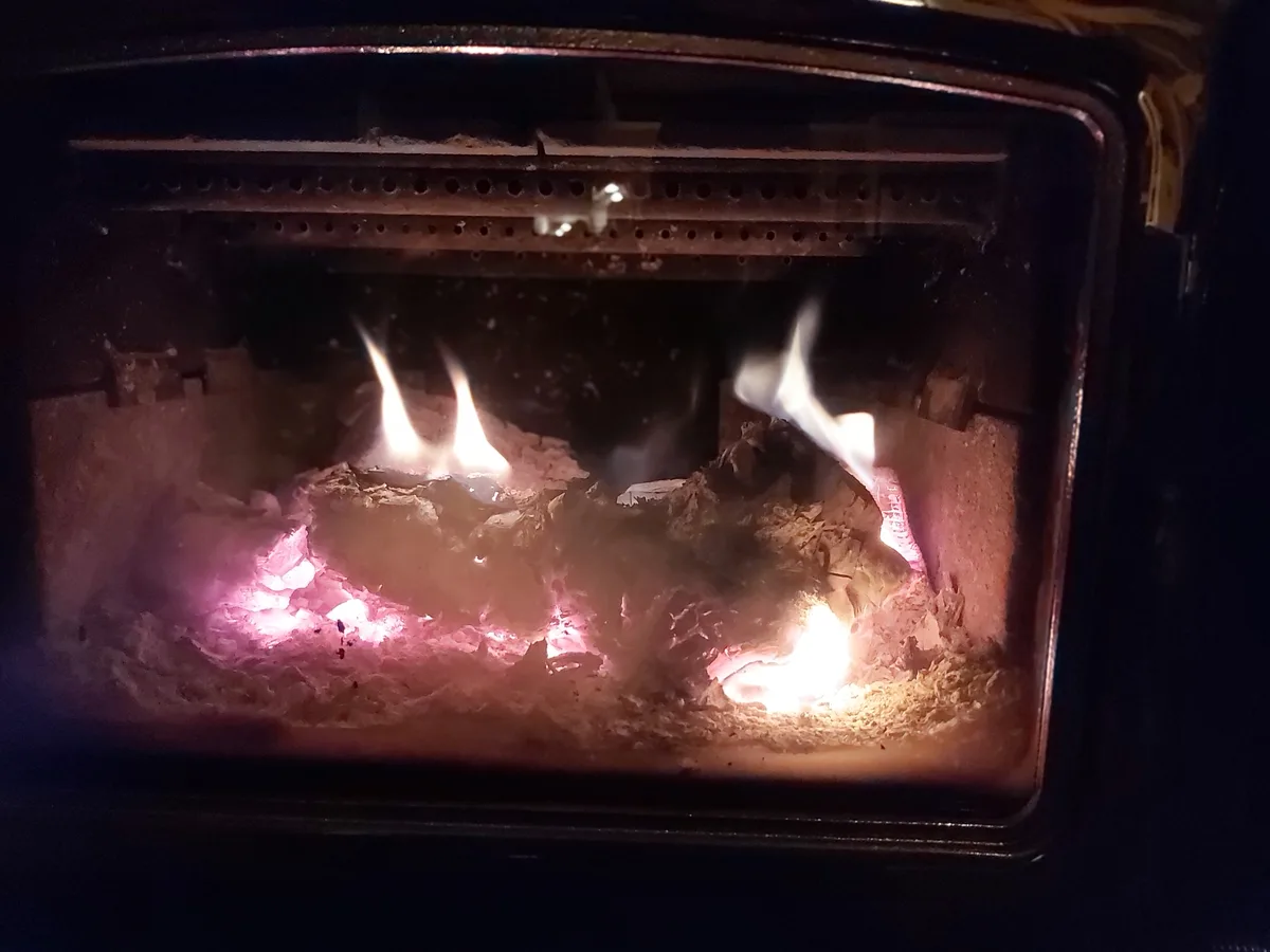 A close-up view of a fireplace or wood stove interior with glowing embers and small flames burning among ashes and charred…