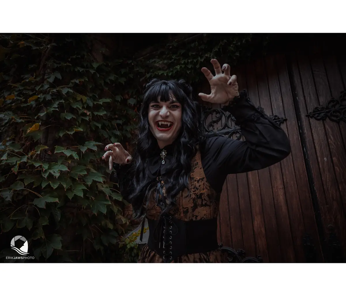 A person dressed as a vampire with long black hair, fangs, and claw-like fingernails, poses dramatically in front of a…