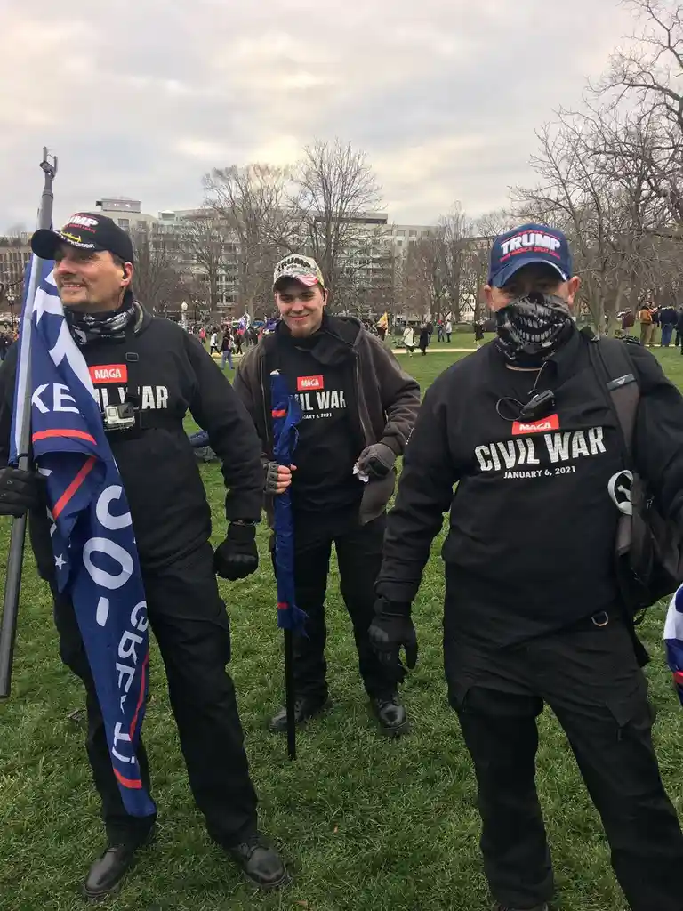Three men in black attire stand on grass wearing 'MAGA' and 'CIVIL WAR JANUARY 6, 2021' shirts, holding flags and wearing…
