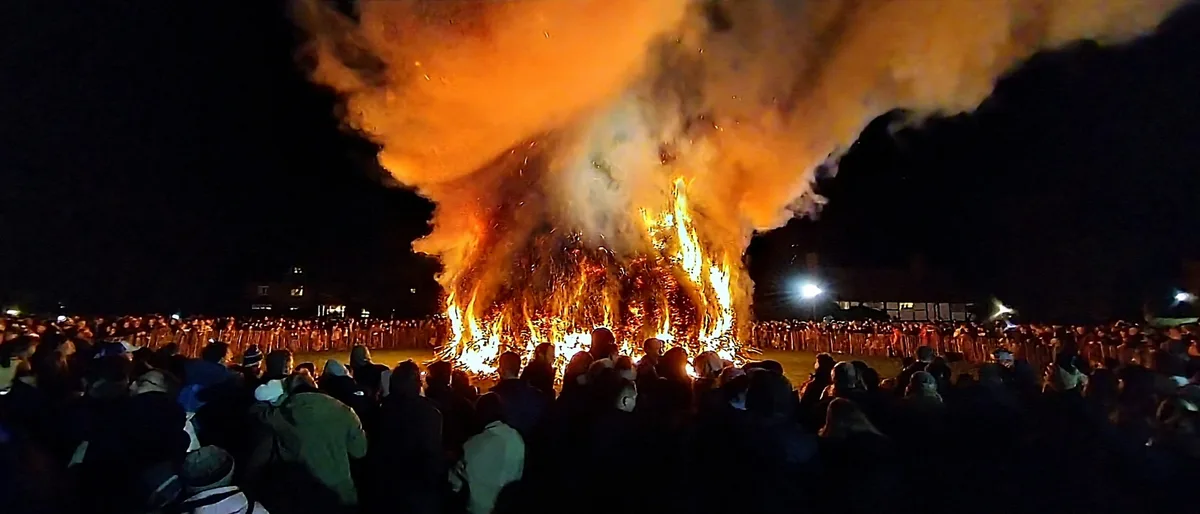 A large crowd watches a massive bonfire erupting at night, flames and smoke billowing upwards. The overlaid text reads 'i'm…