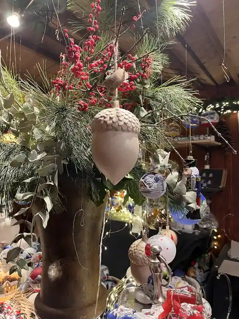 A festive holiday display features a large decorative acorn ornament hanging from a branch with pine needles and red…