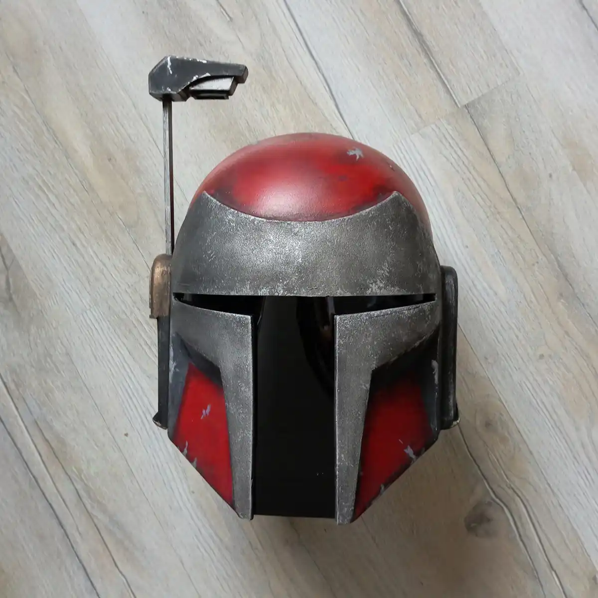 A red and silver Mandalorian helmet, resembling The Mandalorian's iconic design, rests on a light wood floor. The helmet has…