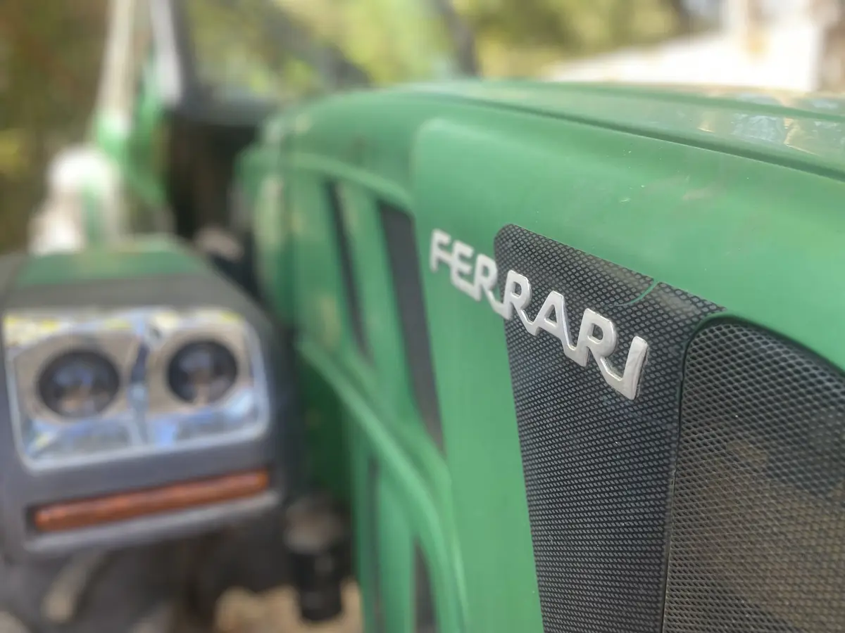 A close-up photo shows the front grille of a green tractor with a silver 'FERRARI' emblem affixed to it. The tractor's…