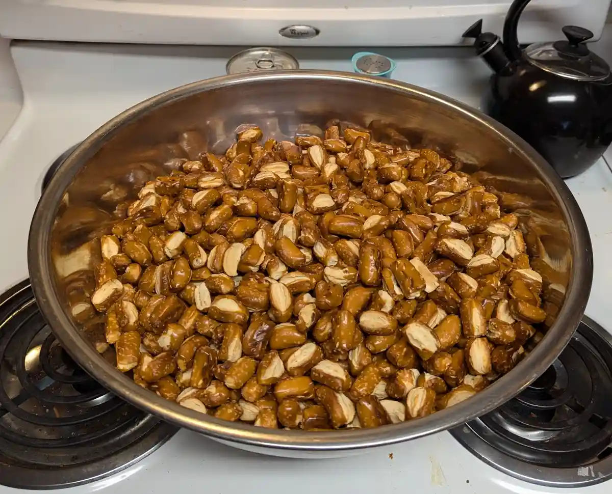 A large metal bowl filled with broken pretzels sits on a white stovetop, with a black kettle and a can visible in the…