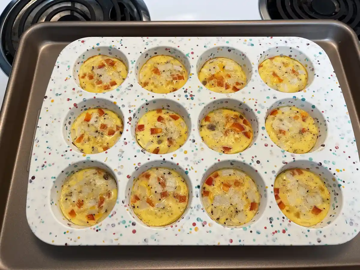 A white silicone muffin tray with colorful speckles sits on a metal baking sheet, containing 12 uncooked egg muffins filled…