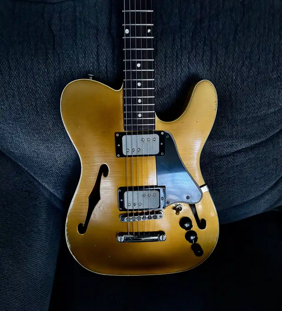A vintage-style hollow-body electric guitar with a sunburst finish and f-holes rests on a dark blue textured couch. The…