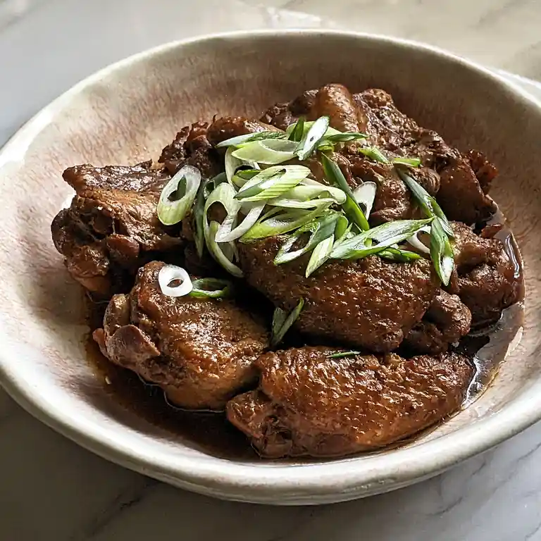 A close-up photo shows a bowl of glossy, dark-brown braised chicken wings garnished with chopped green onions, sitting in a…