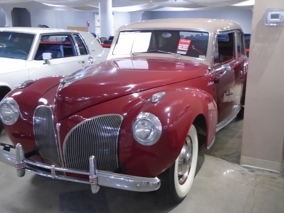 A glossy red vintage car with a beige roof is displayed indoors, likely in a museum or showroom, with a white classic car…