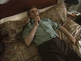 A man lies on a bed with patterned pillows and sheets, holding a microphone to his mouth as if singing or speaking. He wears…