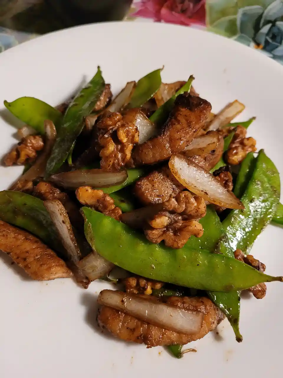 A close-up photo of a white plate holding stir-fried chicken, green snow peas, sliced onions, and toasted walnuts. The food…