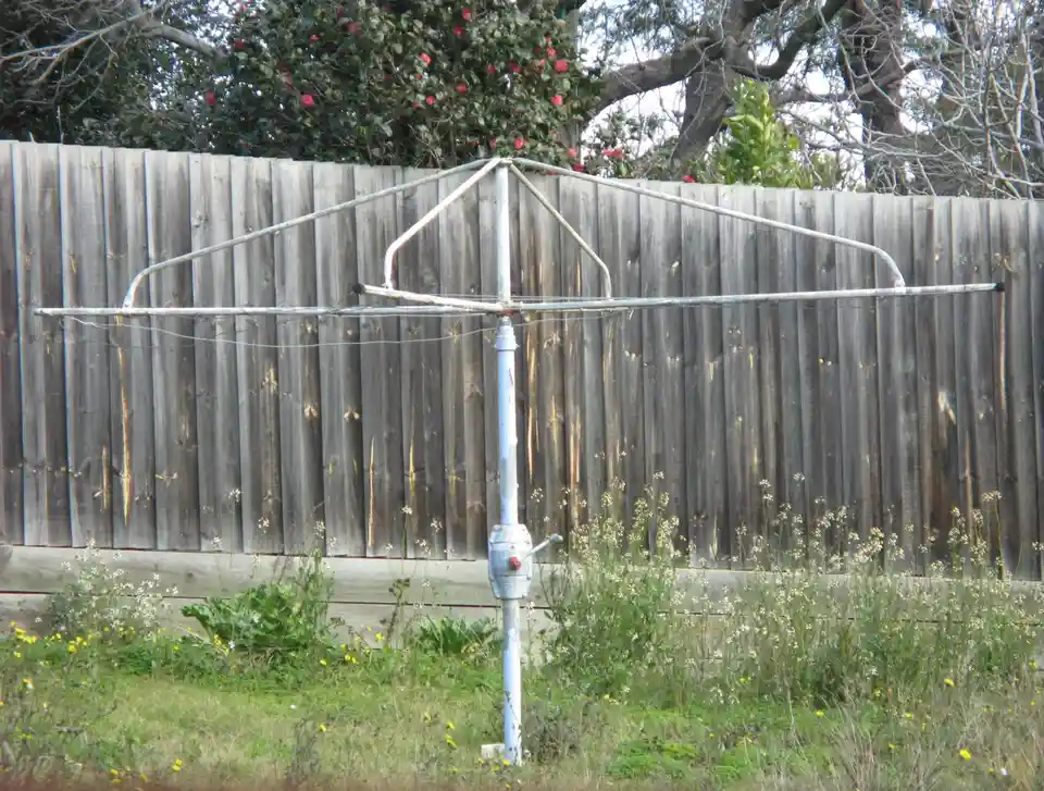 A weathered metal clothesline with a central pole and horizontal bars stands in a grassy yard in front of a wooden fence…