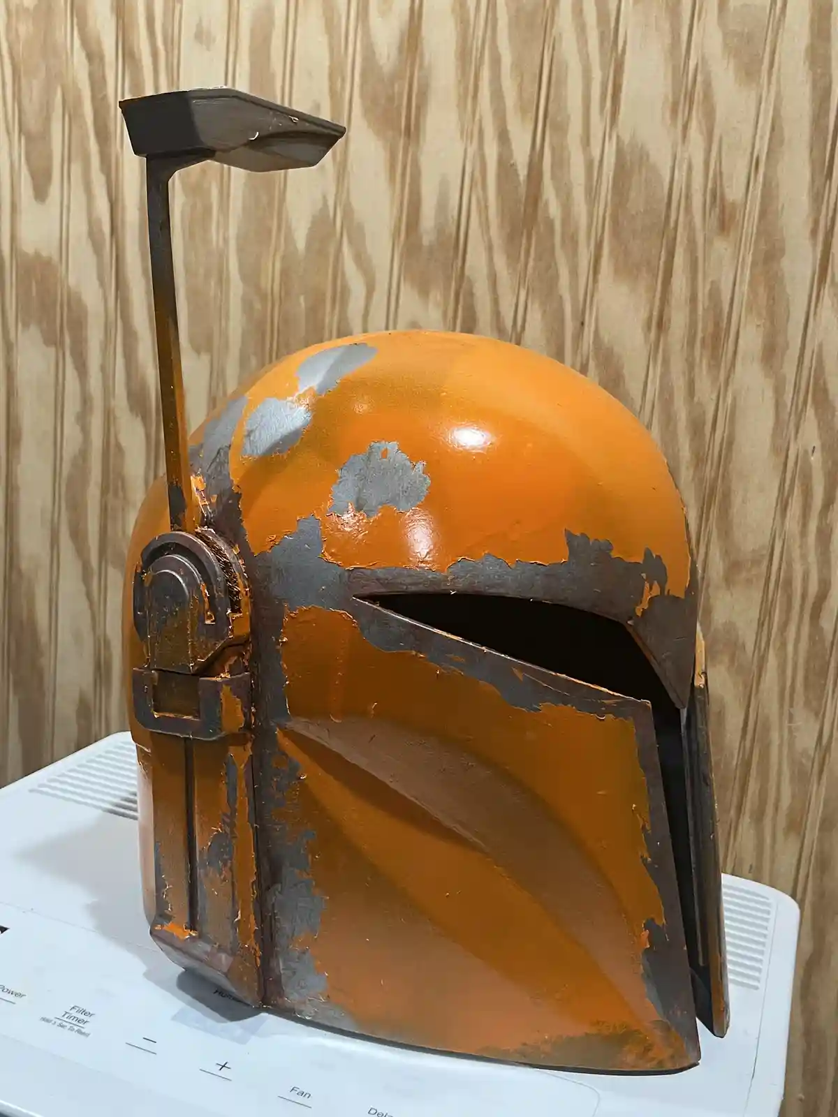A weathered orange and grey Boba Fett helmet with peeling paint sits atop a white appliance, likely a dehumidifier or air…