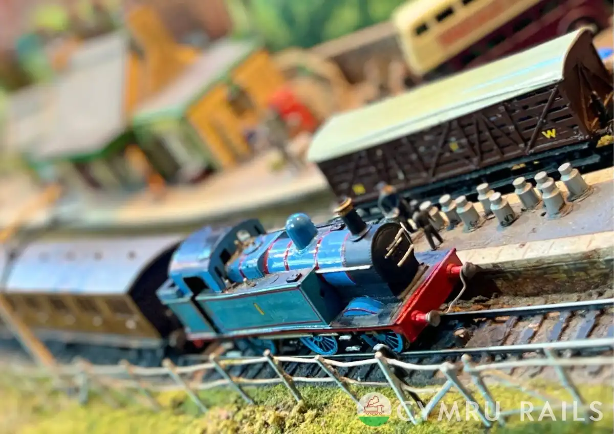 A close-up photo of a blue model train engine on a miniature railway track, pulling a brown freight car, with a blurred…