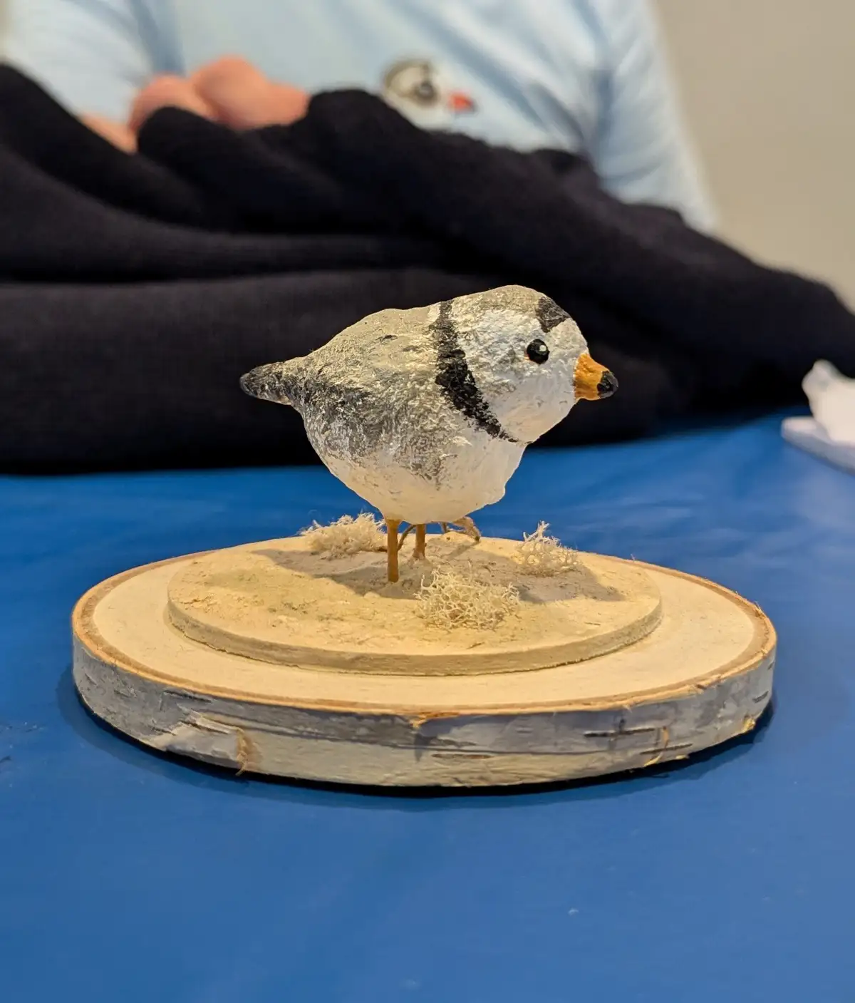 A small, handmade clay bird sculpture with white and black markings, an orange beak, and tiny legs stands on a circular…