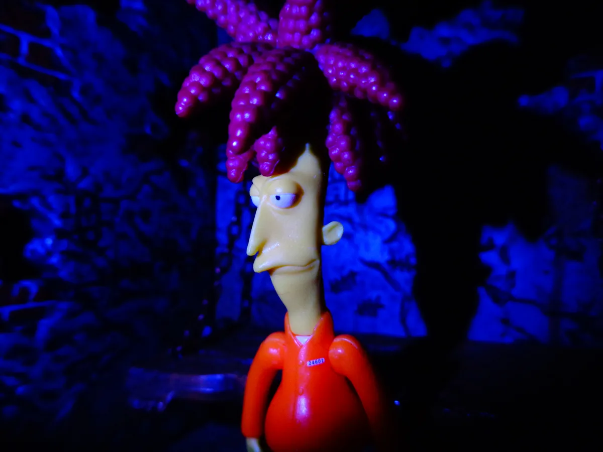 A plastic figurine of the animated character Fry from Futurama, wearing an orange jumpsuit with a visible tag reading…