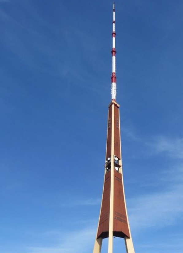 A tall, slender tower with a brown, tapering base and a red-and-white striped antenna array against a clear blue sky. The…