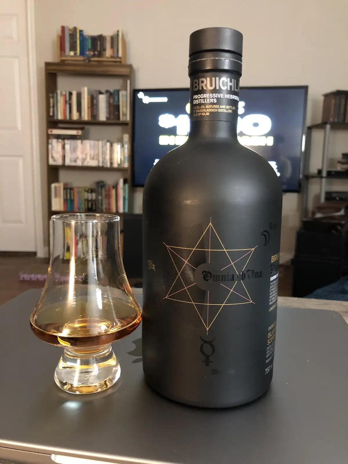 A black bottle of Bruichladdich 'Omnia ab Una' whisky sits next to a tasting glass with amber liquid on a dark table, with a…