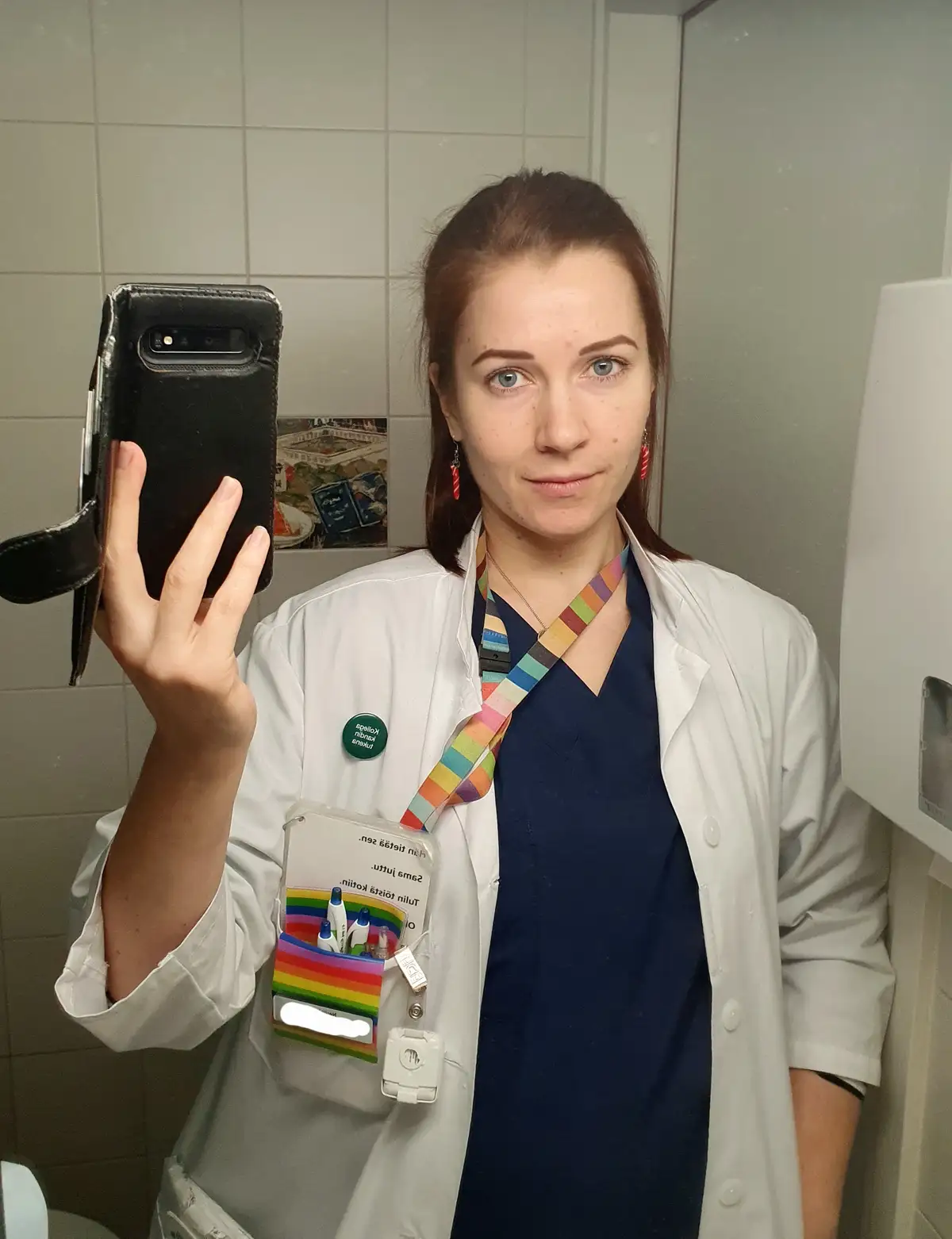A woman in a white lab coat and blue scrubs takes a mirror selfie in a tiled bathroom, holding a black phone. Her lanyard…