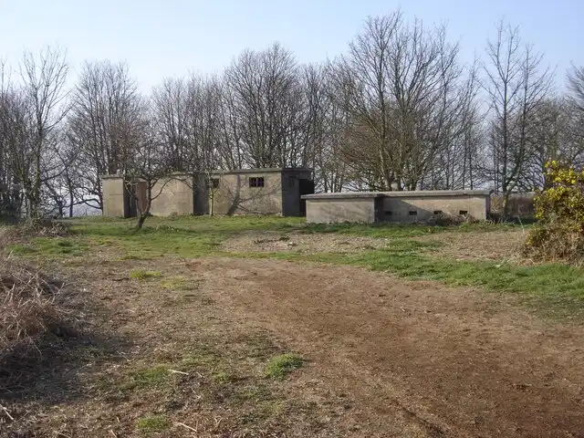 An outdoor photo shows a dirt path leading to several dilapidated, concrete block buildings surrounded by bare trees and…