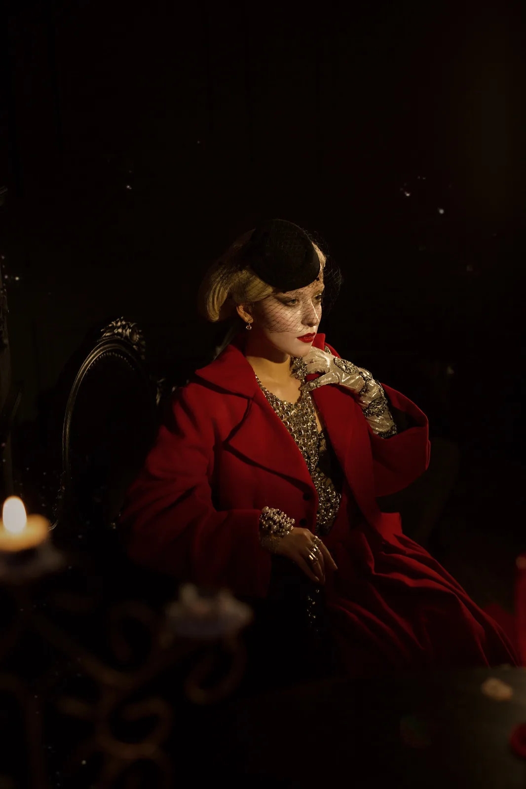 A woman with blonde hair and a black netted hat sits in a dark, candlelit room wearing a red coat over a jeweled dress and…