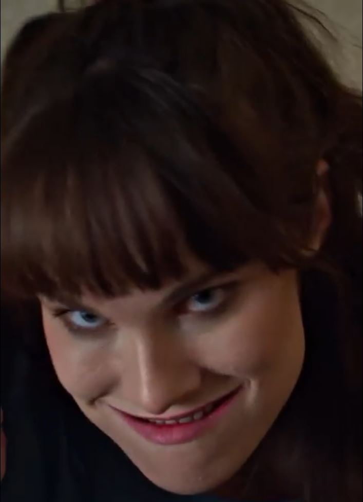 A close-up shot of a woman with brown hair and bangs, looking down with a smug, knowing smile and narrowed eyes. The…