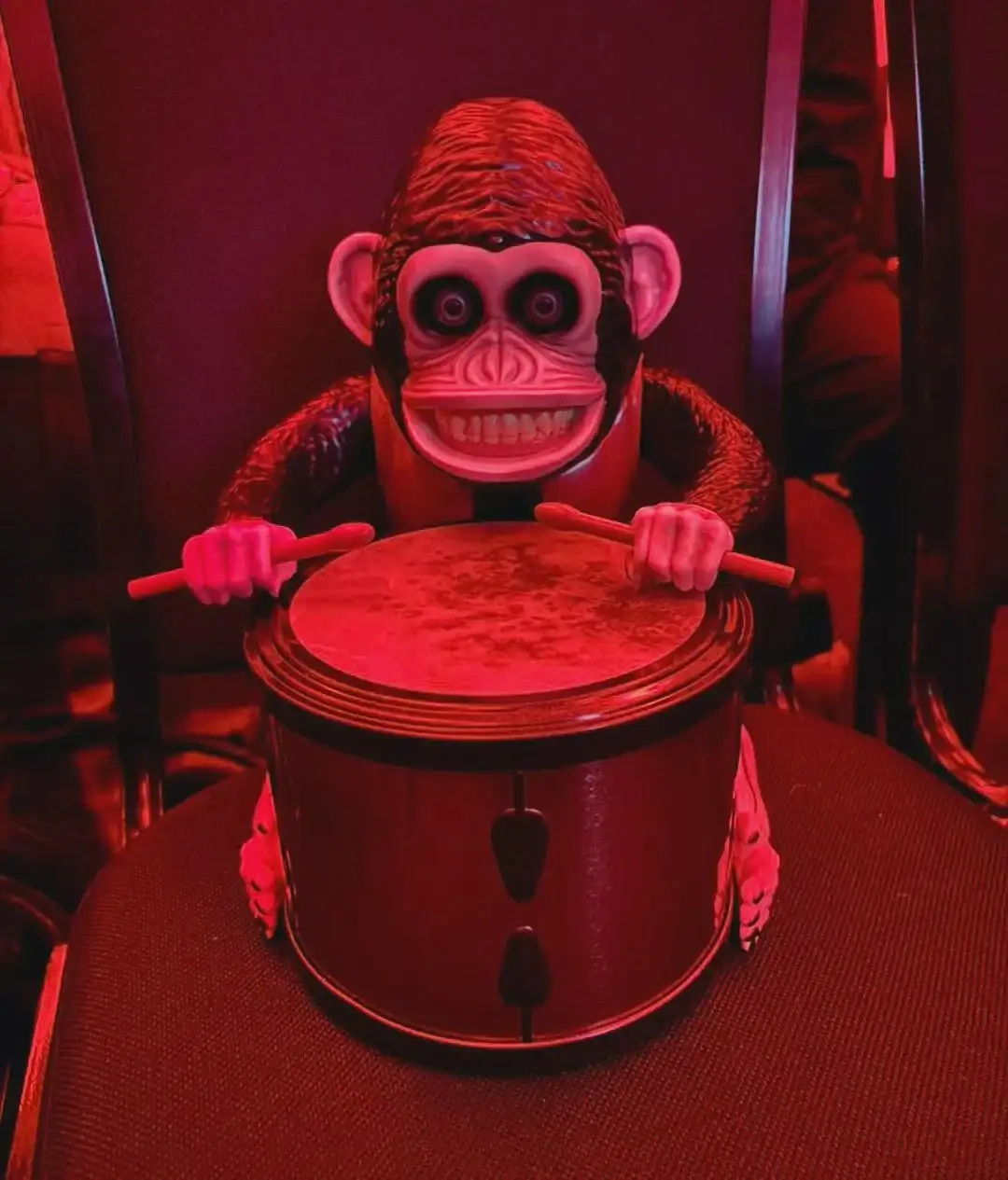 A plastic monkey toy with a wide, toothy grin and large eyes sits on a chair, holding drumsticks over a small drum. The…