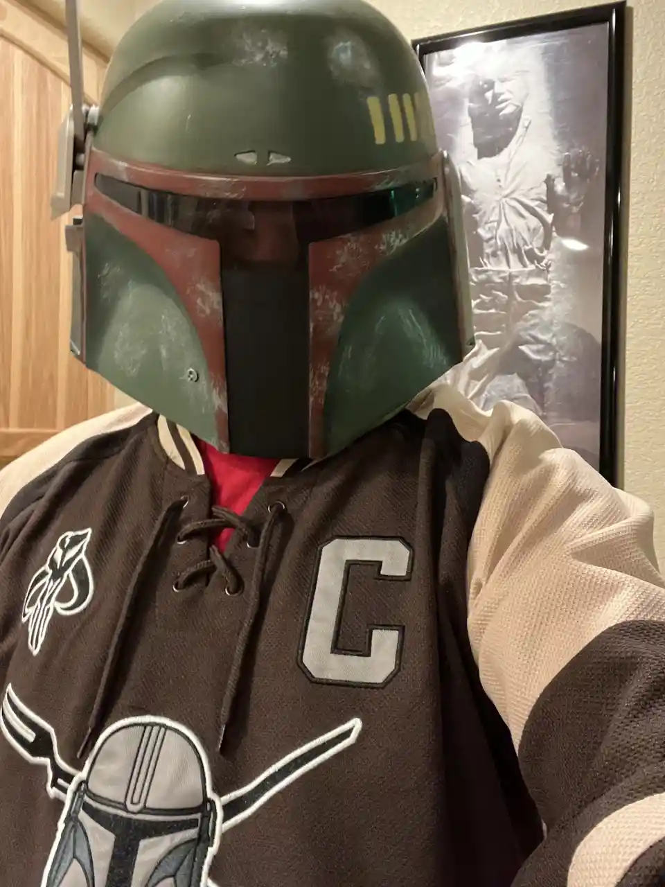A person wearing a weathered Boba Fett helmet and a brown hockey jersey with a large 'C' on the chest and a Mandalorian…