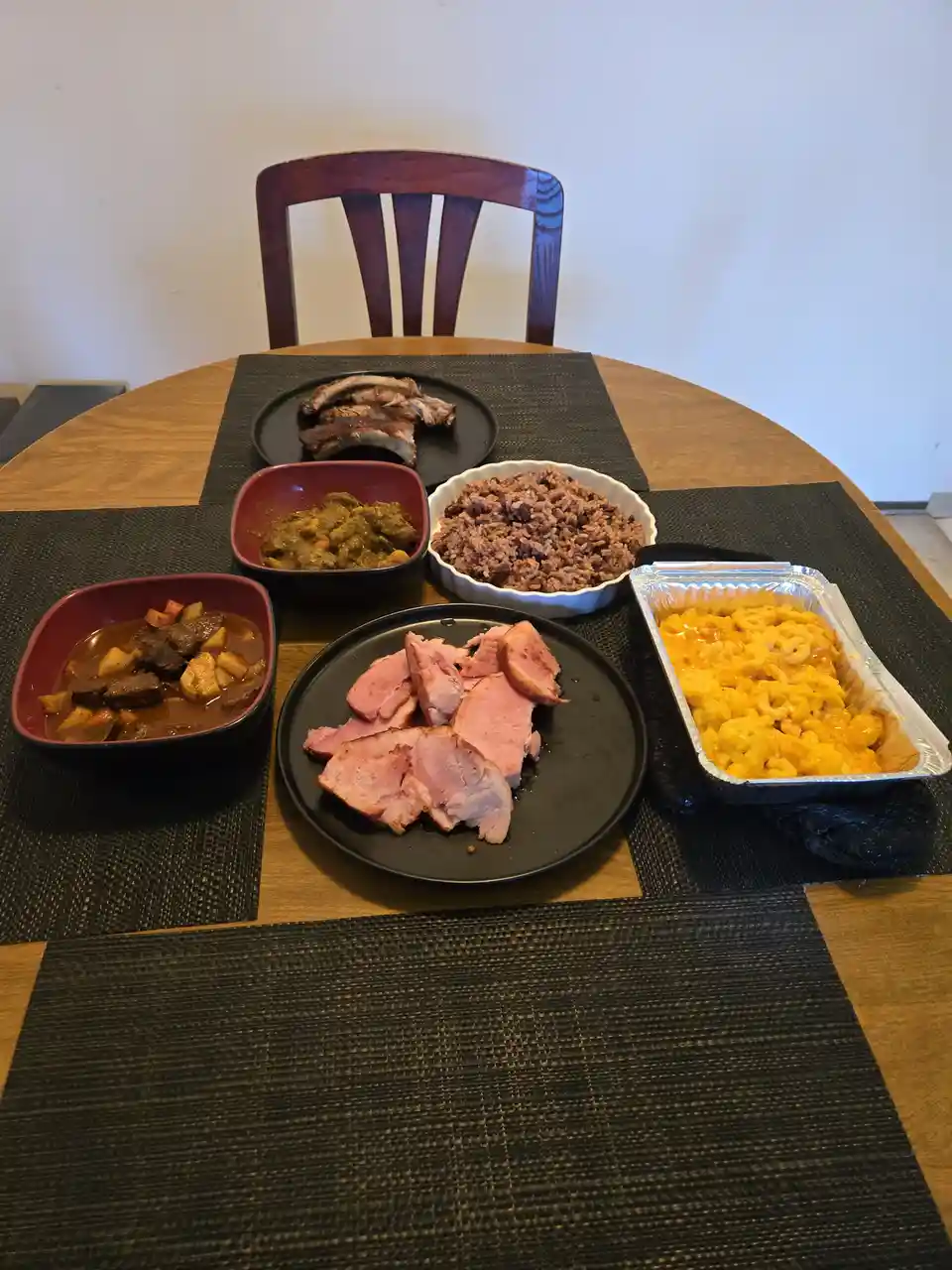 A wooden dining table with a dark woven placemat holds six dishes: ribs, sliced ham, macaroni and cheese in a foil pan…