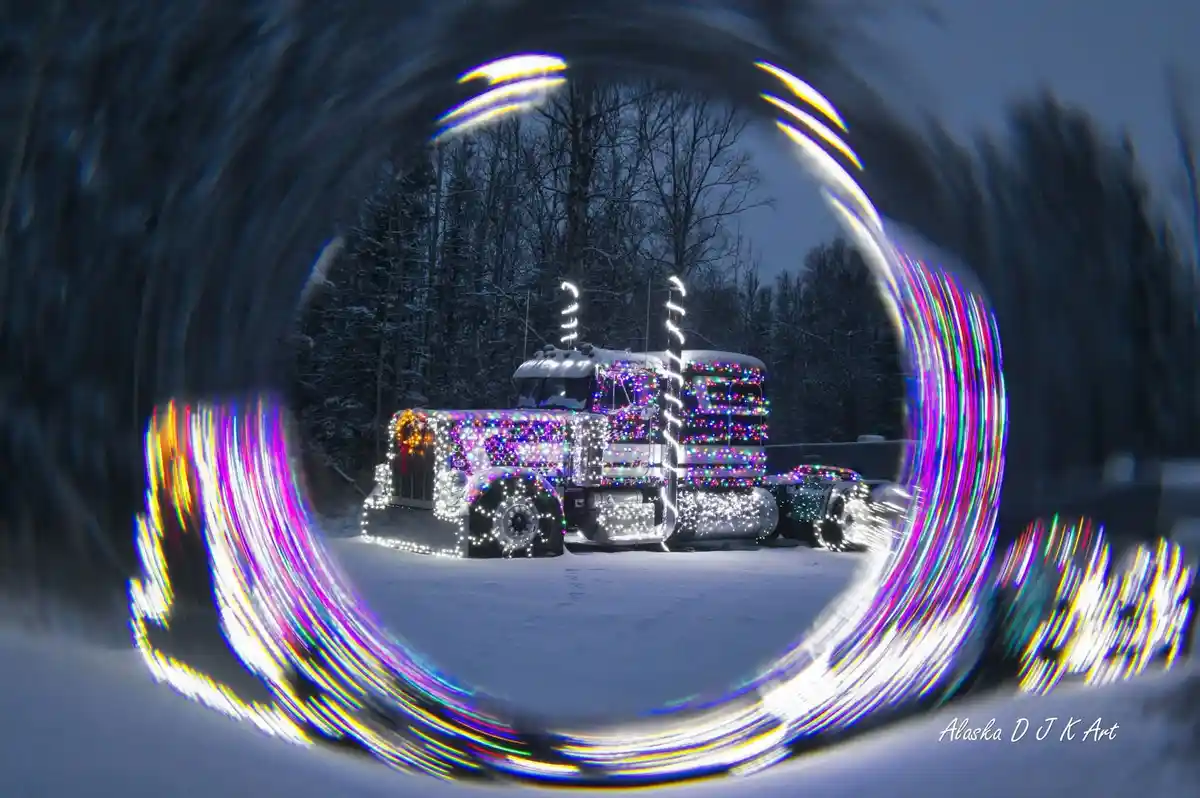 A large, snow-covered semi-truck is heavily decorated with colorful Christmas lights, viewed through a fisheye lens that…