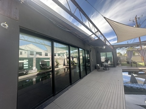 A modern outdoor patio with large glass sliding doors reflecting a pool and house, under a shade sail and metal frame…