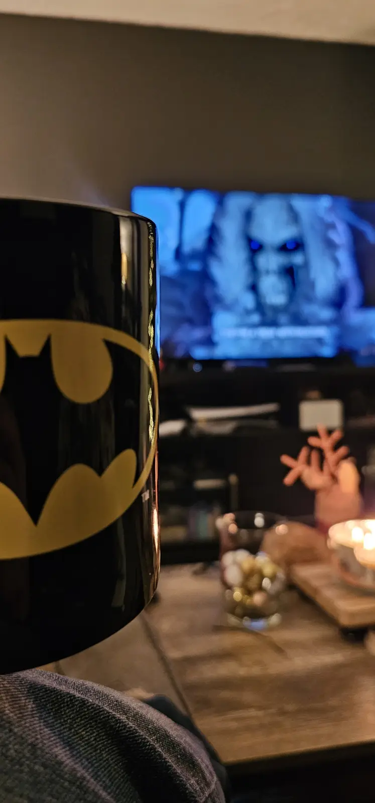 A close-up of a black Batman mug with a yellow bat symbol is in the foreground, obscuring a blurry background showing a TV…