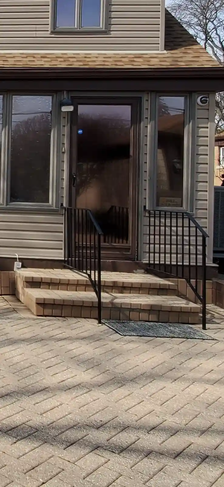 A photo of a residential front door with brick steps and black railings, featuring a small sign in the right window that…