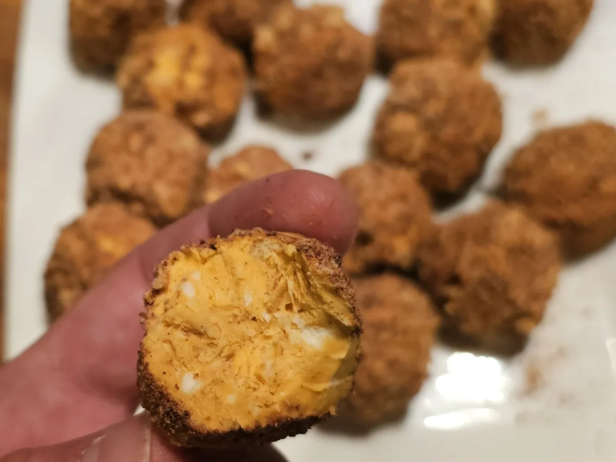 A hand holds a halved, golden-brown fried ball revealing a creamy, orange-yellow interior with white flecks, likely cheese…