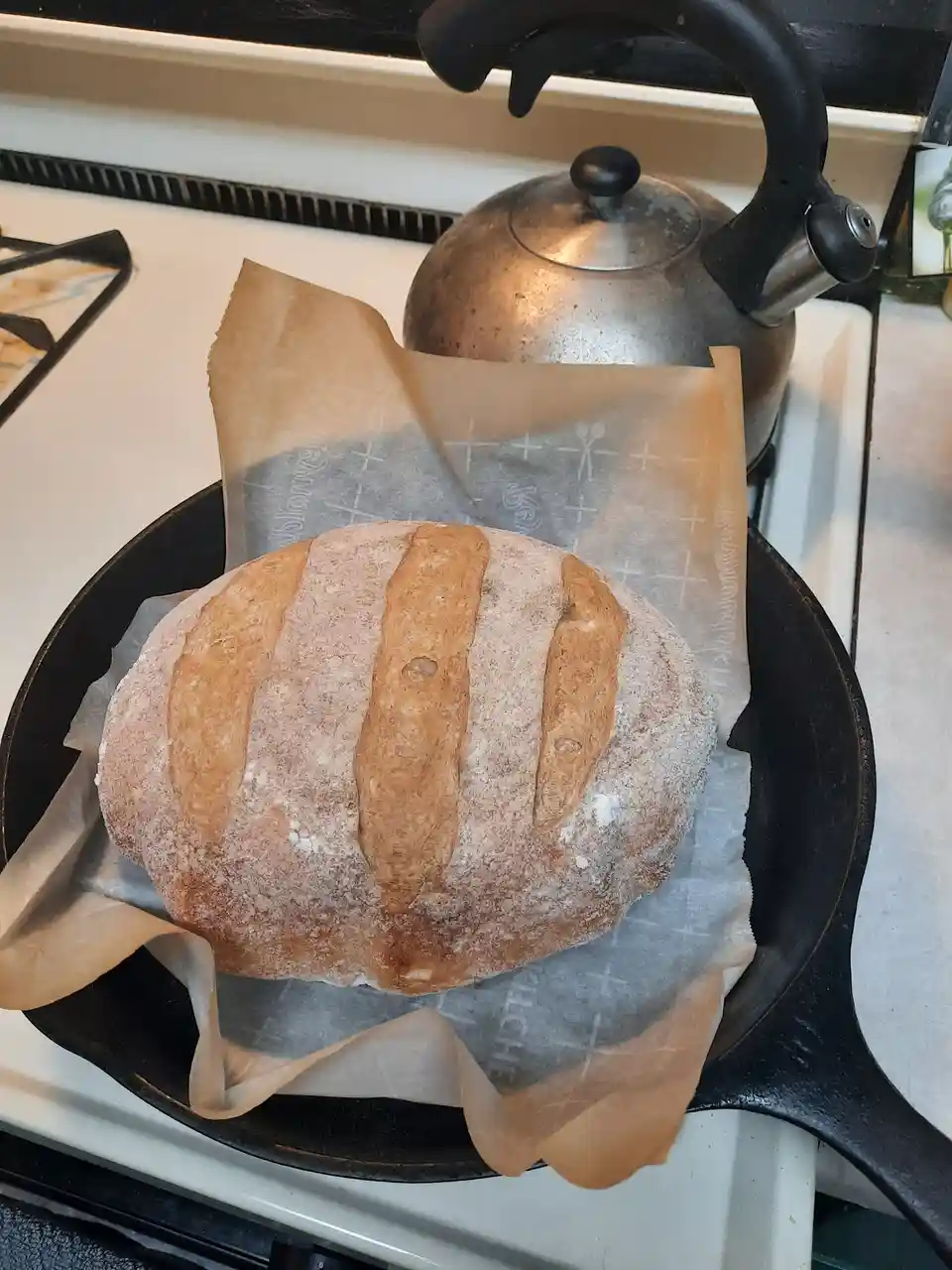A round, rustic loaf of bread with three scored slashes sits on parchment paper inside a black cast-iron skillet on a…