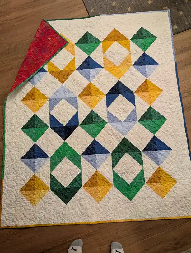 A colorful handmade quilt with a geometric pattern of green, blue, and yellow diamonds on a cream background is laid out on…