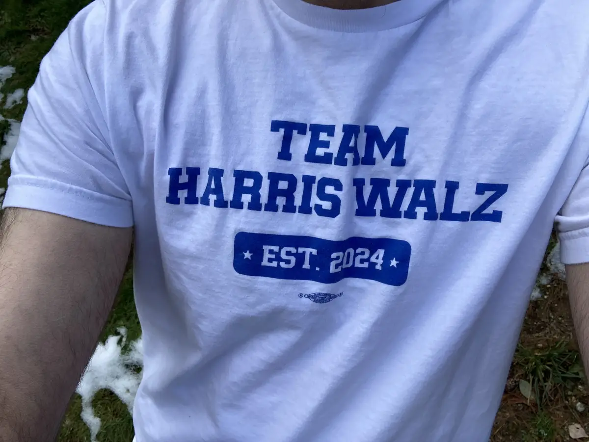 A close-up photo shows a person wearing a white t-shirt with blue text that reads 'TEAM HARRIS WALZ' and 'EST. 2024' inside…