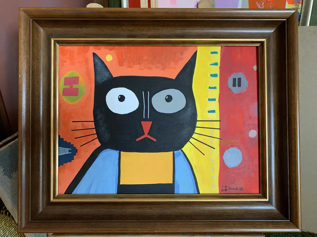 A framed painting of a stylized black cat with large white eyes and a red nose, set against a colorful abstract background…