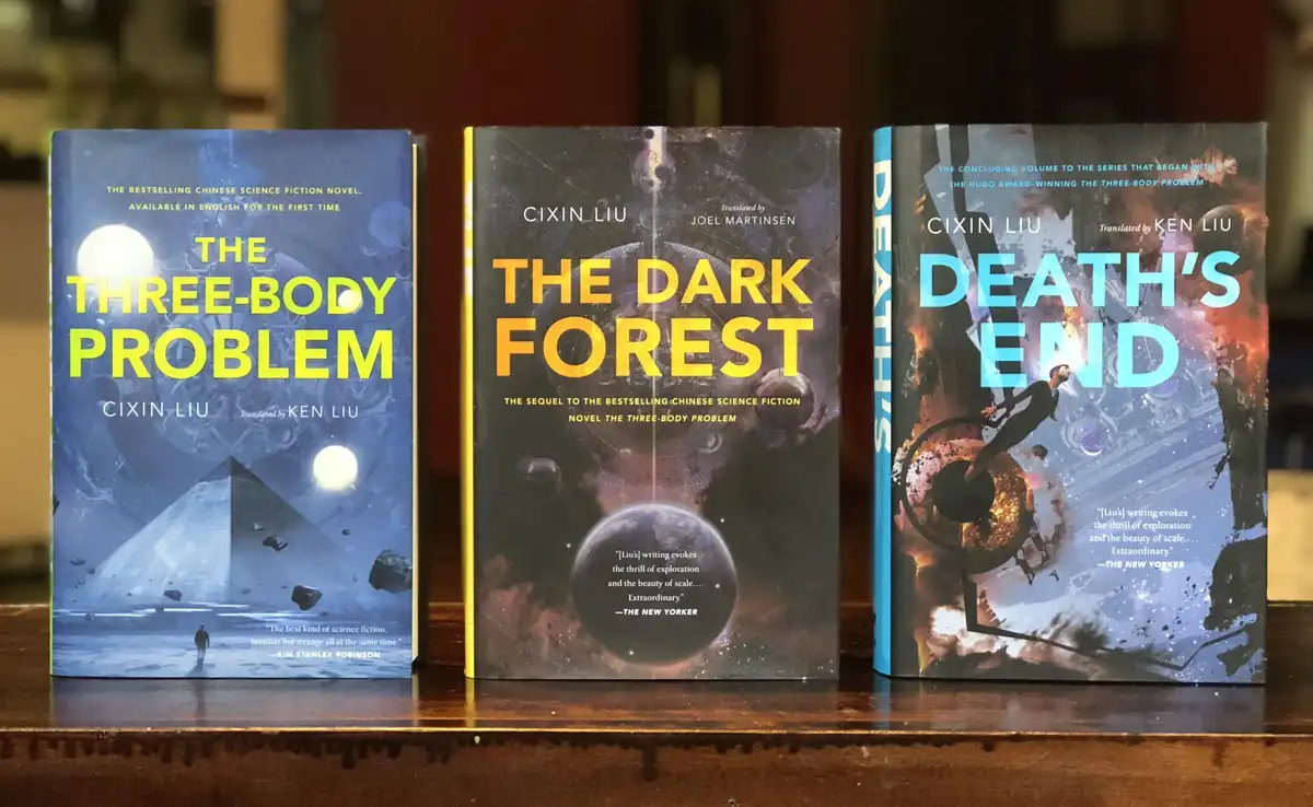 Three books by Cixin Liu are displayed side-by-side on a wooden surface: 'The Three-Body Problem', 'The Dark Forest', and…