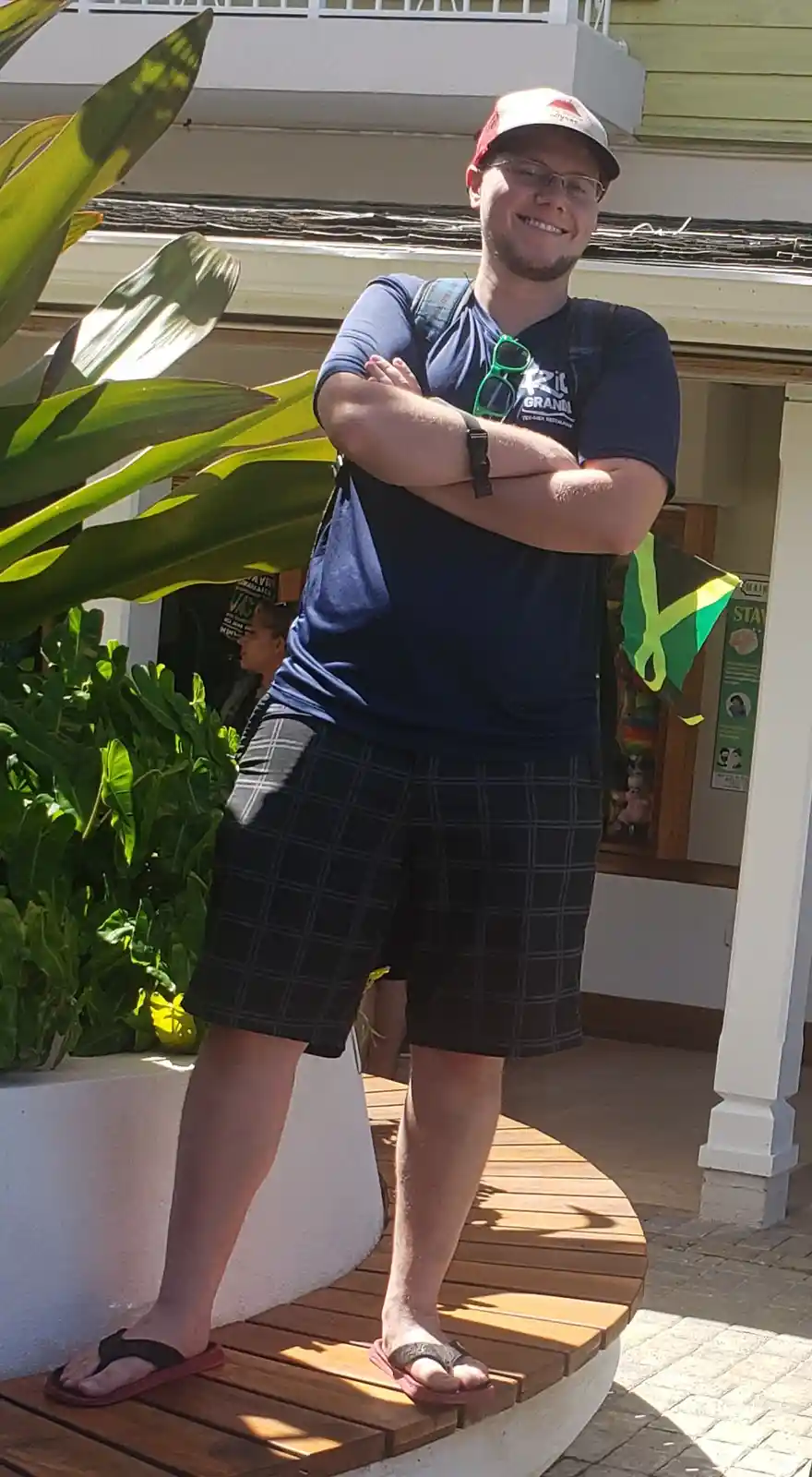 A man with glasses and a cap stands confidently with arms crossed on a wooden platform outdoors, smiling at the camera. He…