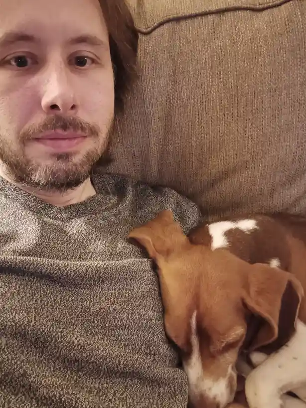 A man with a beard and brown hair lies on a brown couch, wearing a grey sweater, looking directly at the camera with a…