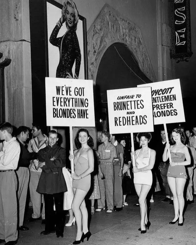 A black-and-white photo from the 1950s shows three women in vintage swimwear holding protest signs outside a theater with a…
