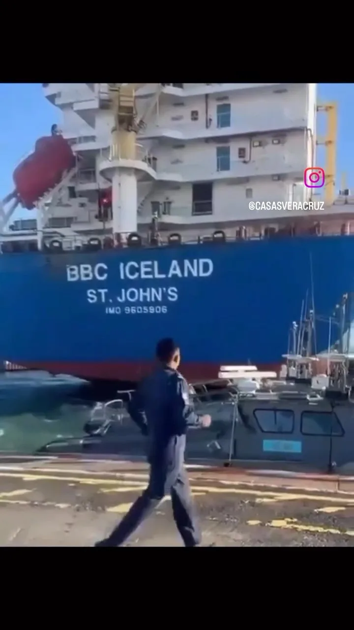 A man in a dark uniform walks away from the camera on a dock, with a large blue cargo ship behind him. The ship's hull…