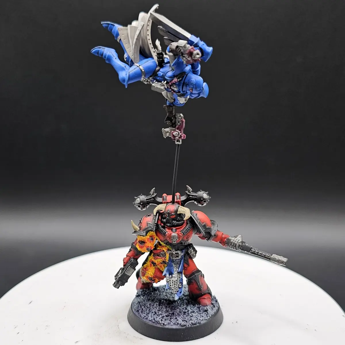A red and black painted miniature of a heavily armored warrior stands on a circular base, holding a weapon and looking…